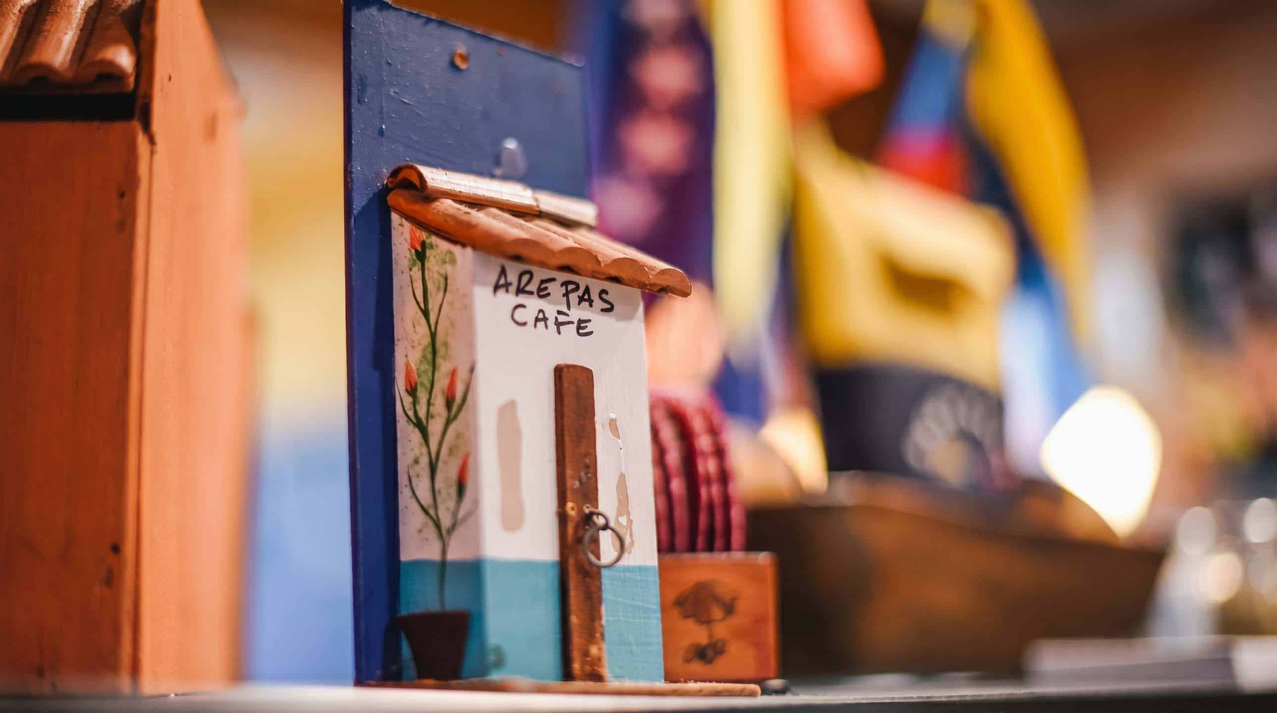 Hand-painted miniature Arepas Café sign on display inside the restaurant, with Venezuelan flag in the background.